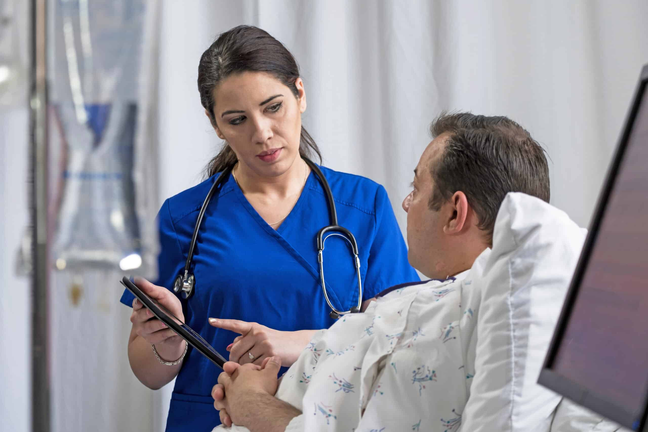 Nurse reviewing medical information with a patient at bedside using a tablet in a hospital setting.