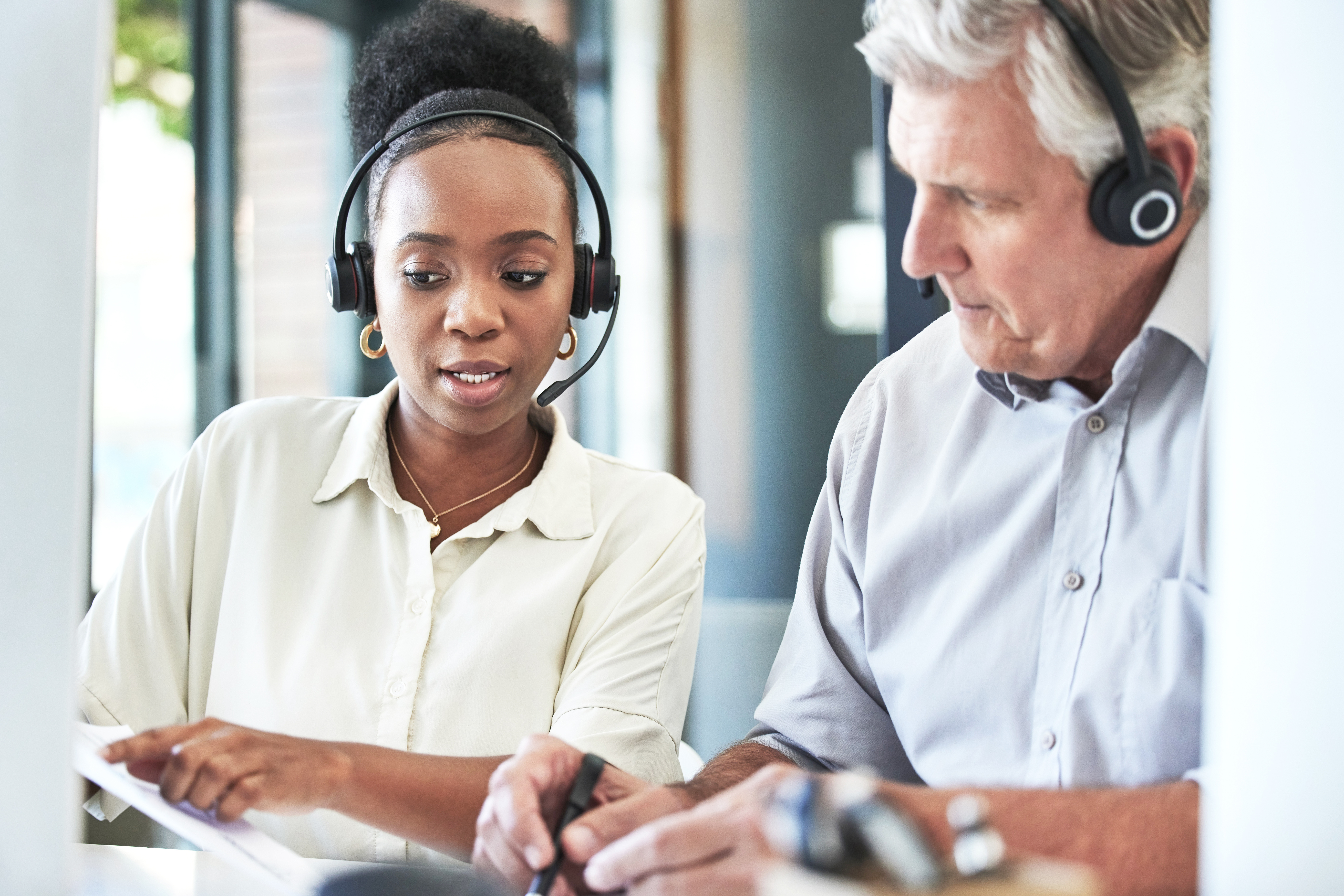 Two professionals wearing headsets collaborate while reviewing paperwork together in an office.