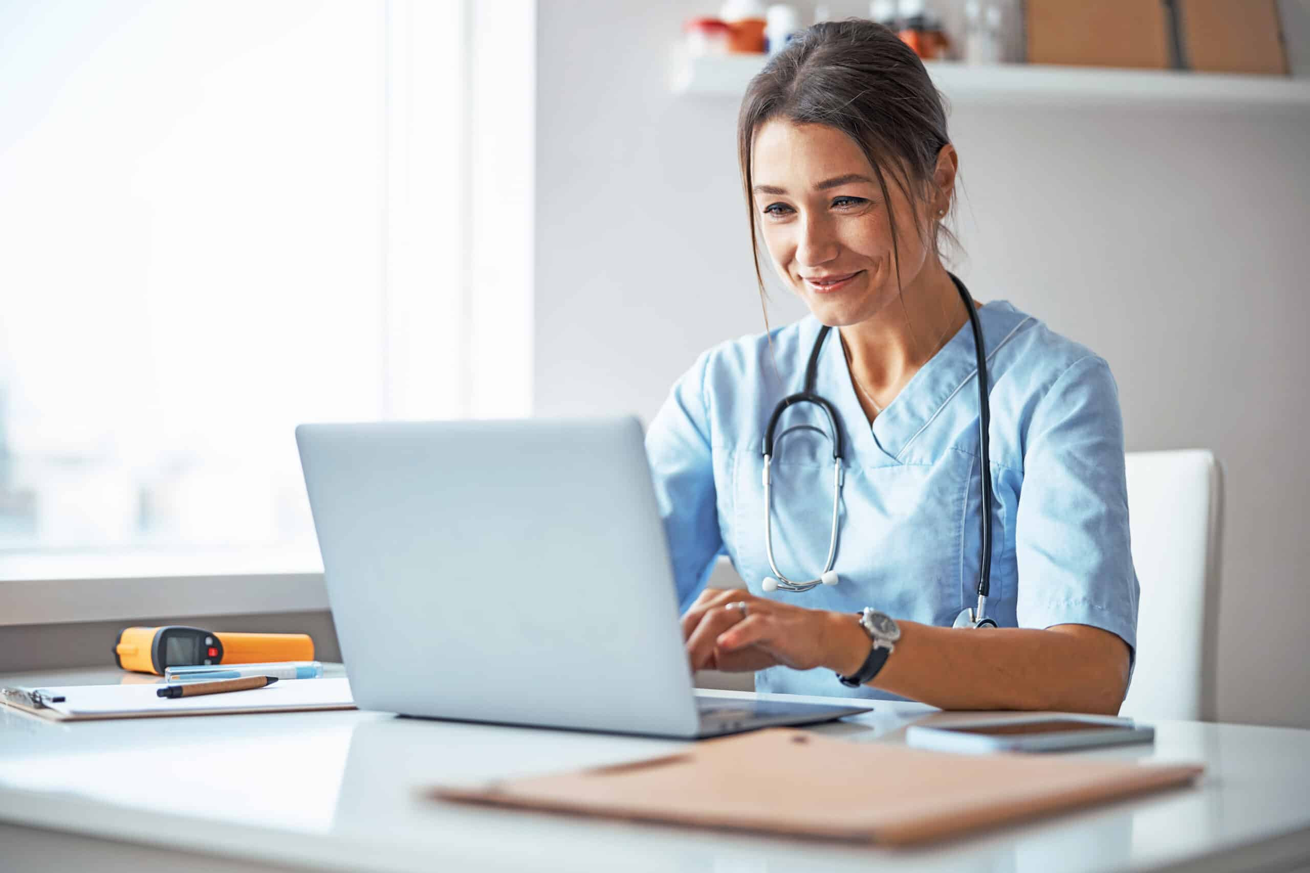 Healthcare professional in scrubs using a laptop, representing digital health systems and clinical workflow efficiency.