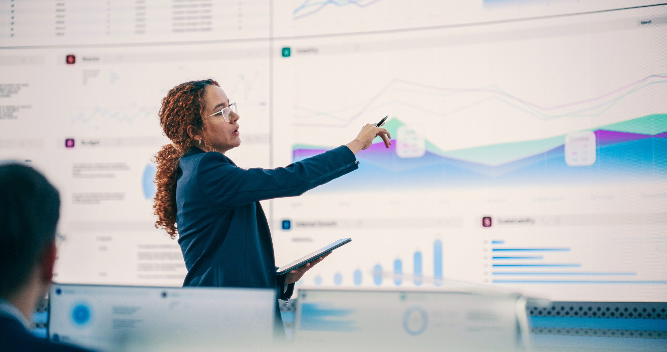 Hispanic Female Senior Data Scientist Reviewing Reports Of Risk Management Department On Big Digital Screen In Monitoring Room. Diverse Consulting Company Employees Working Behind Desktop Computers.