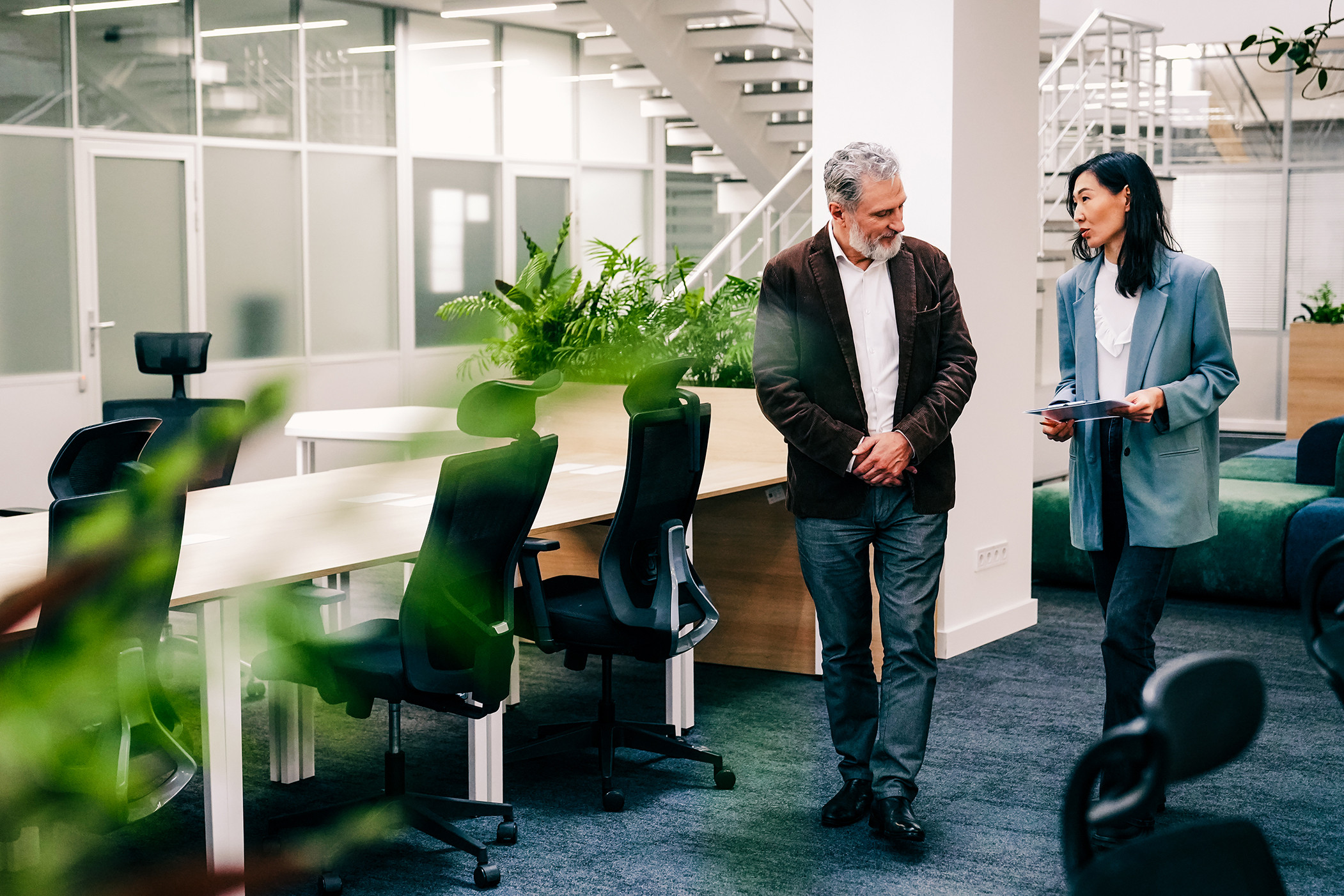 A middle-aged man and woman are walking and discussing documents in a modern open-plan office. They are business partners engaged in a professional conversation near workstations.