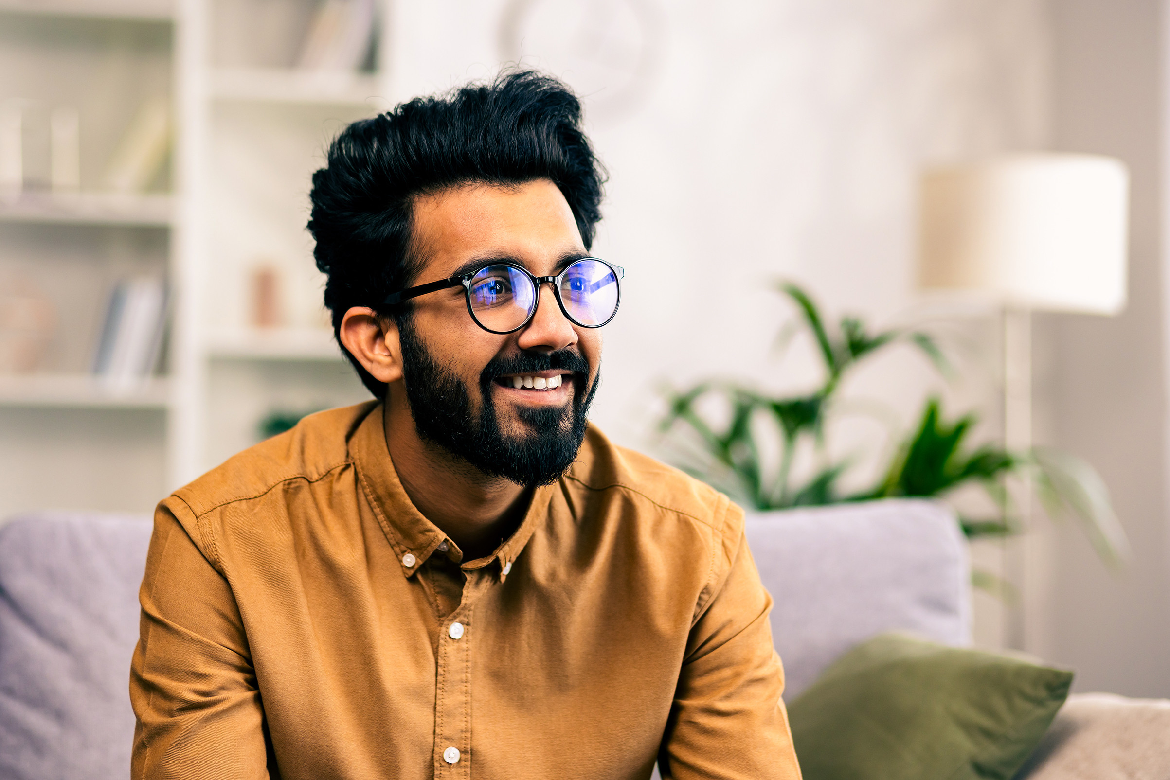 Close-up photo of a young Indian man in glasses sitting on the couch at home and looking to the side with a smile.