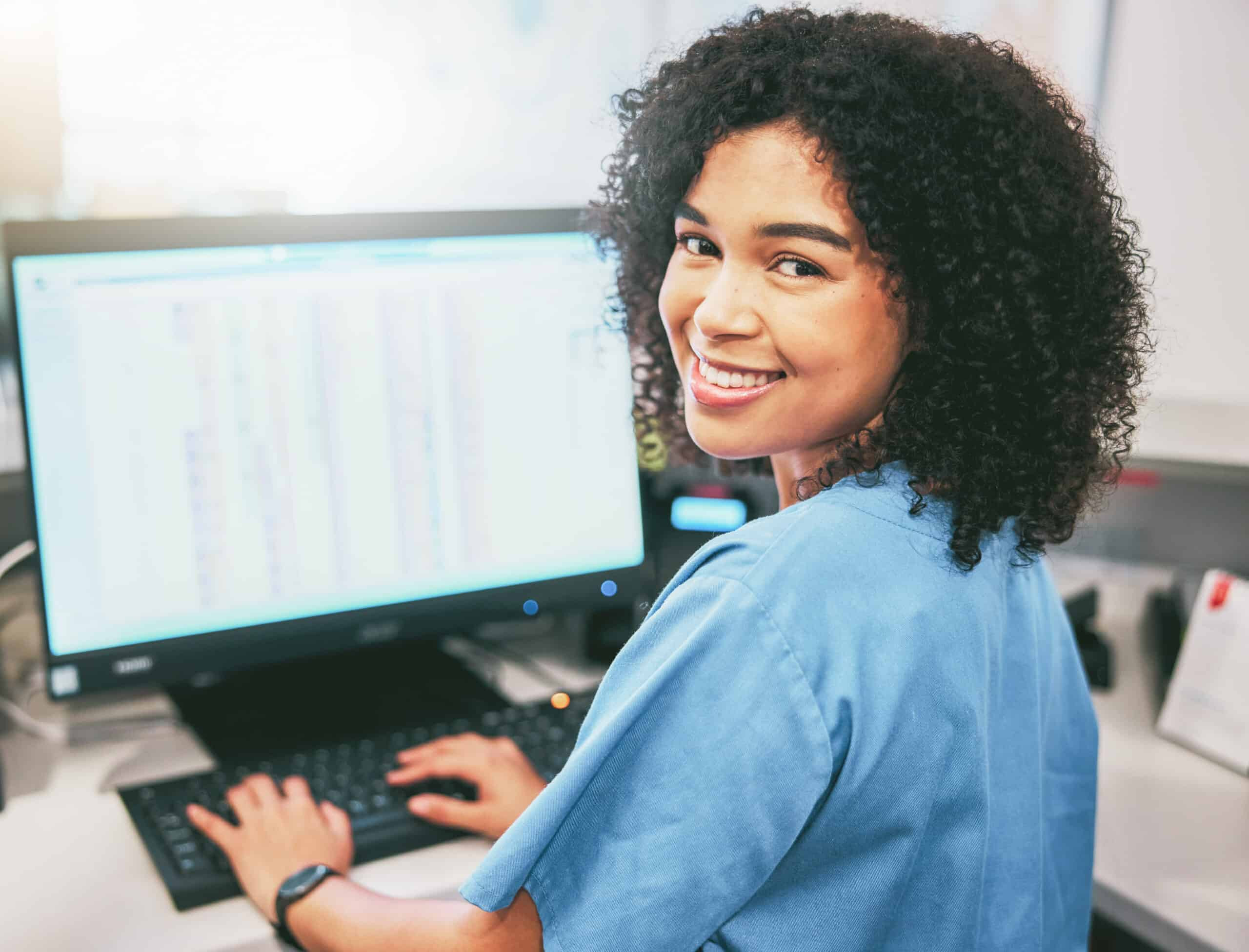 Healthcare professional working at a computer, smiling while reviewing patient or administrative data.