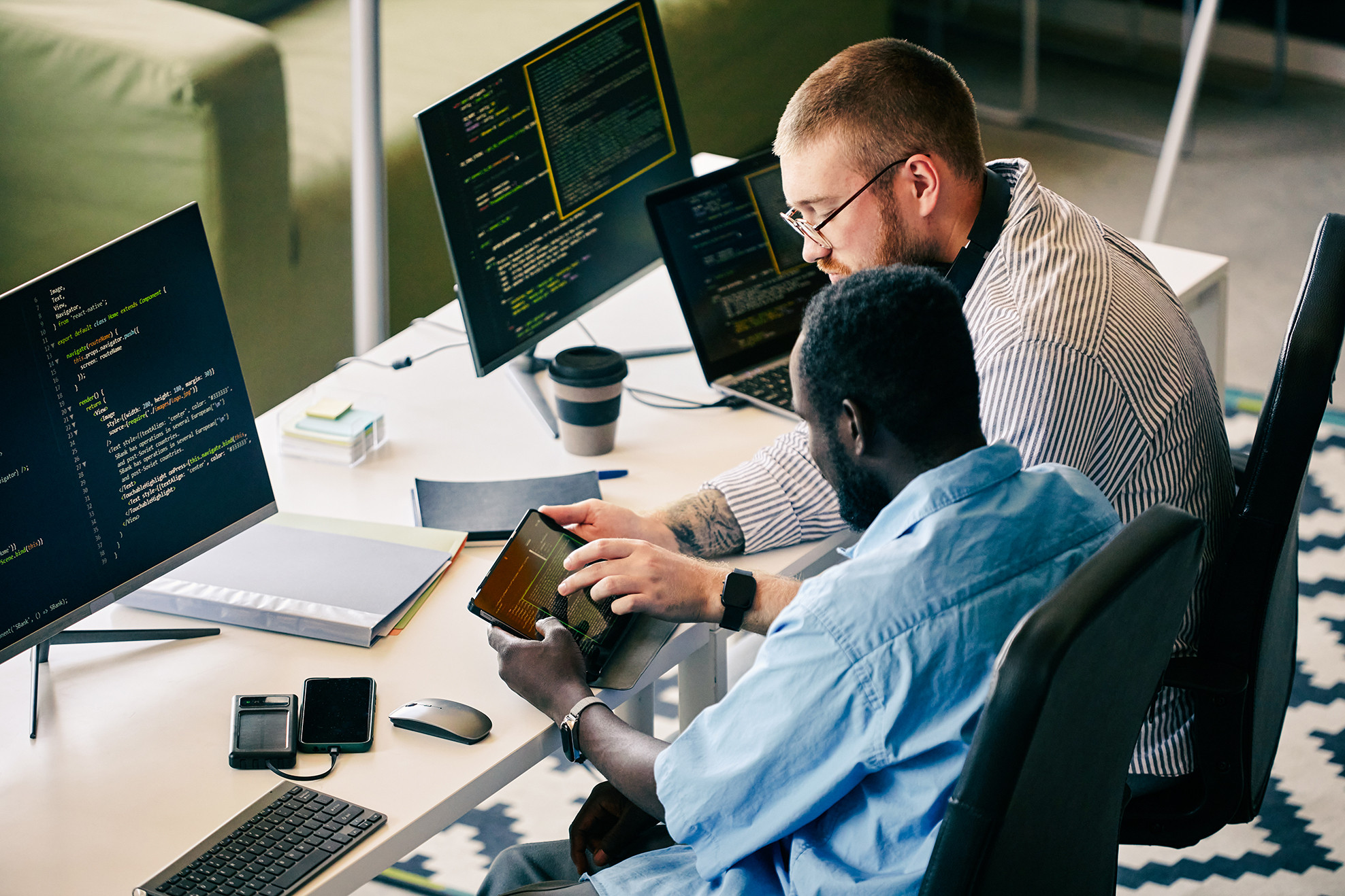 Two colleagues working together on software development in modern tech workspace, both focused on coding and using digital tools. One person is showing information on a tablet