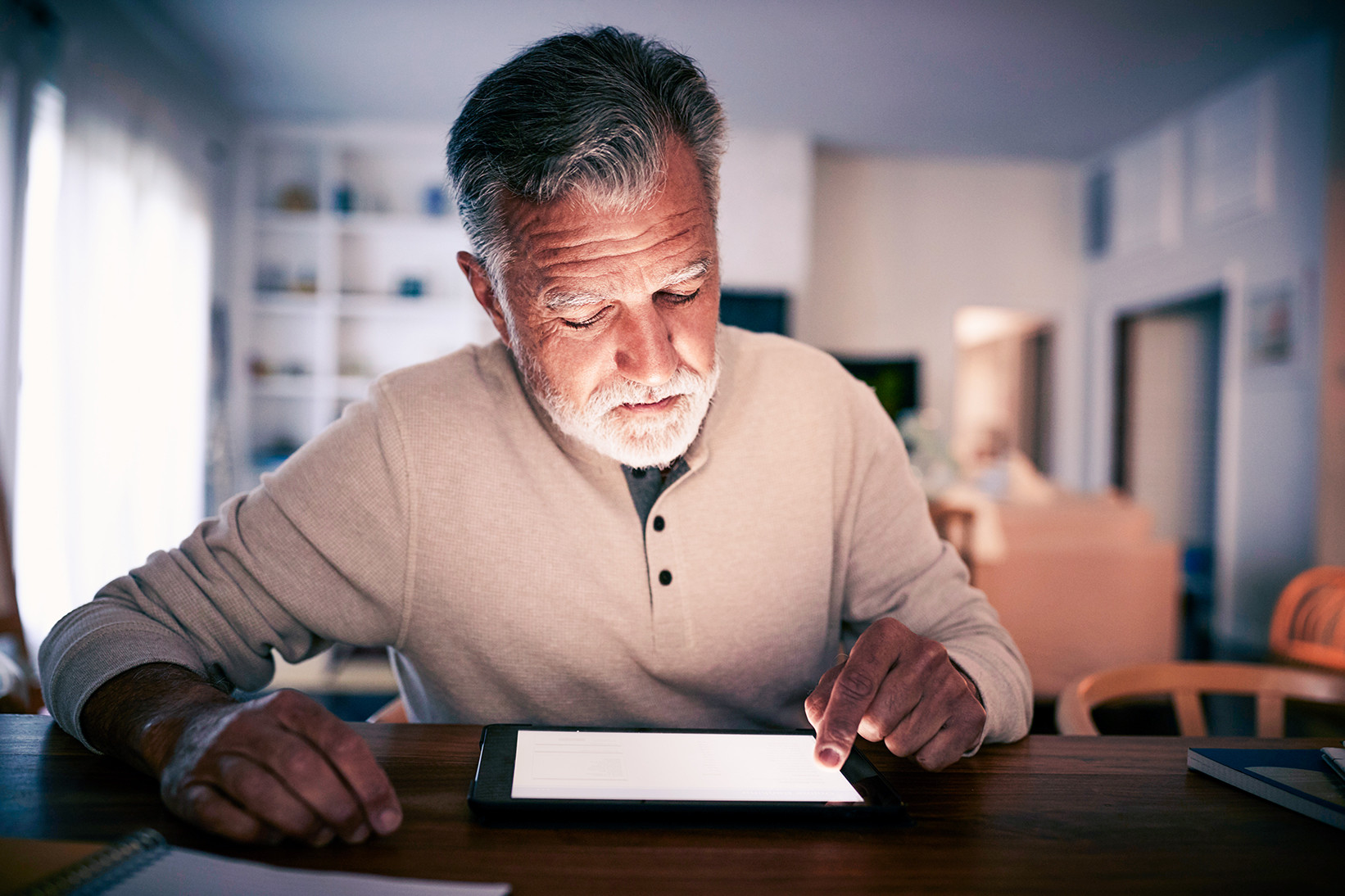 Senior Hispanic man sitting at a table using a tablet computer at home in the evening, close up