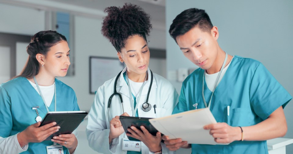 Healthcare professionals reviewing patient records together on tablets and clipboard in a clinical setting.