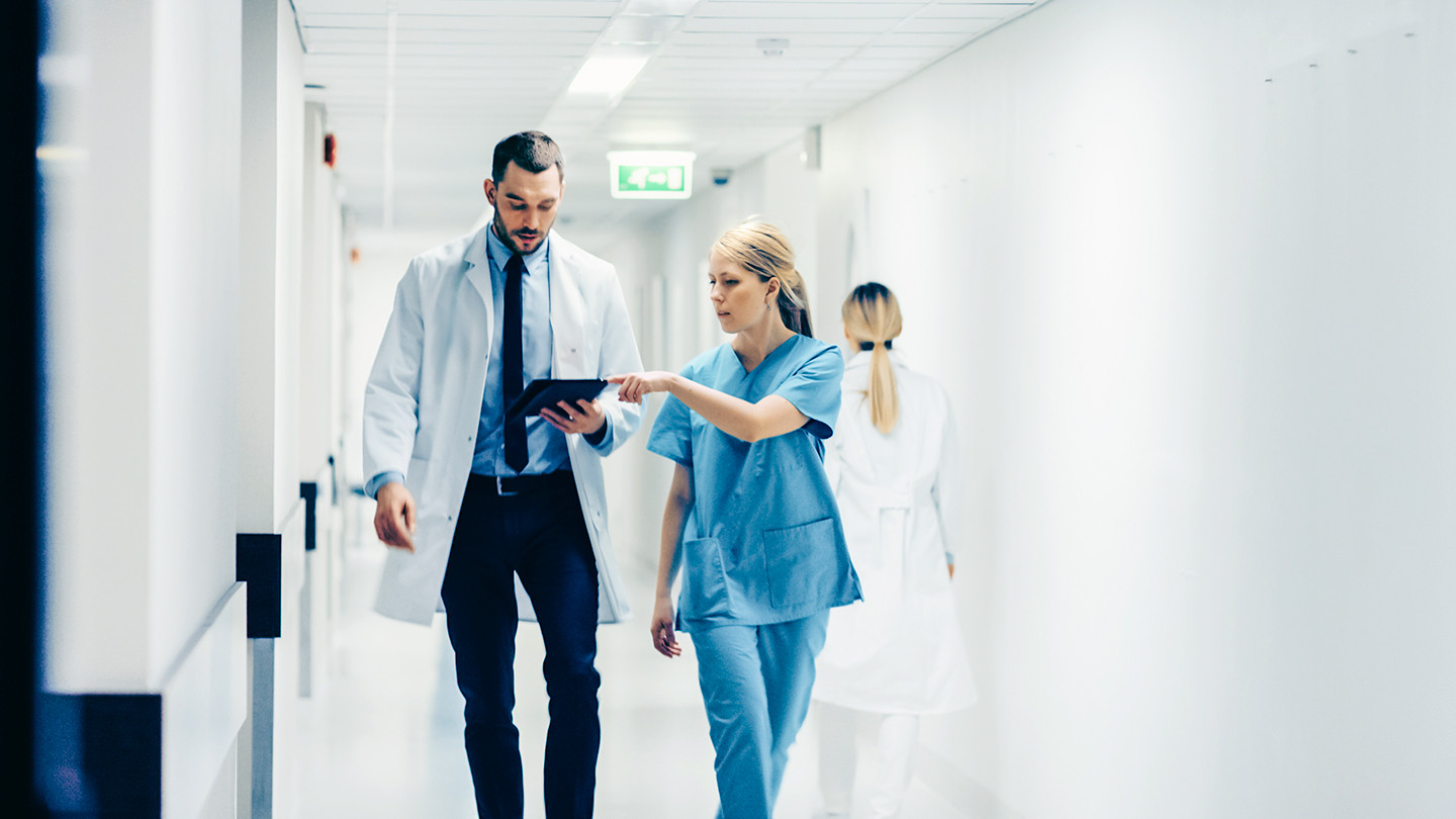 Female Surgeon and Doctor Walk Through Hospital Hallway, They Consult Digital Tablet Computer while Talking about Patient's Health. Modern Bright Hospital with Professional Staff.