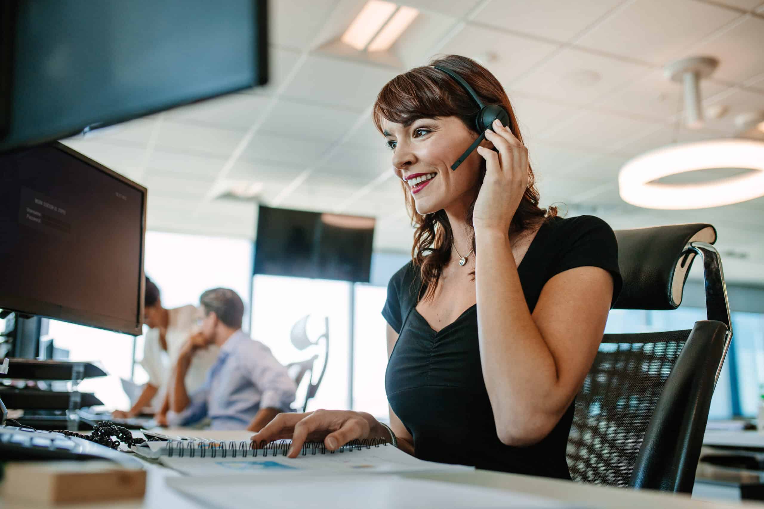 Woman smiles answering the phone on a headset in a call center setting|||