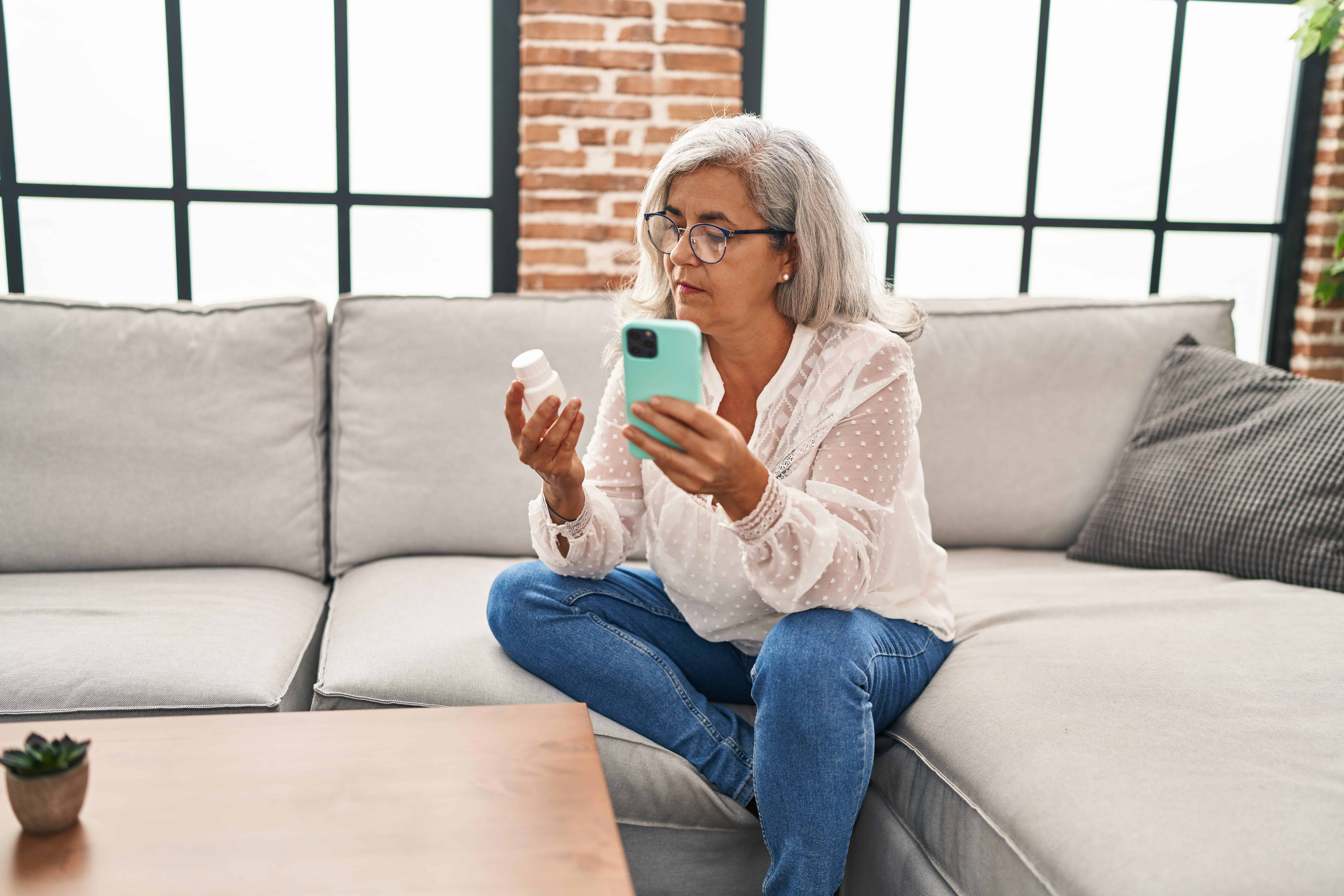 Middle age woman using smartphone holding pills at home